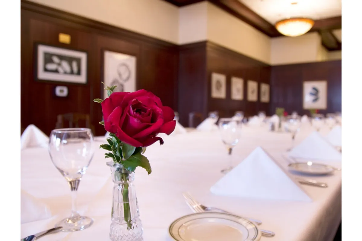 A sophisticated dining setting with a neatly arranged table, a red rose in a vase, and framed art on the walls. The ambiance is elegant yet welcoming—perfect for business meals or celebratory gatherings.