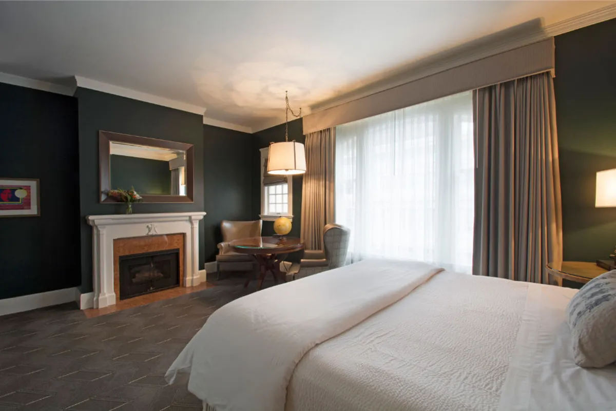 The image showcases an elegant hotel room with a plush king-sized bed, a glowing fireplace, an inviting seating area, and large windows letting in abundant natural light. The decor is sophisticated and welcoming, blending modern luxury with cozy touches. Guests can imagine unwinding in the soft seating, enjoying a book or a warm drink by the fire, while the city of Portland beckons just outside.