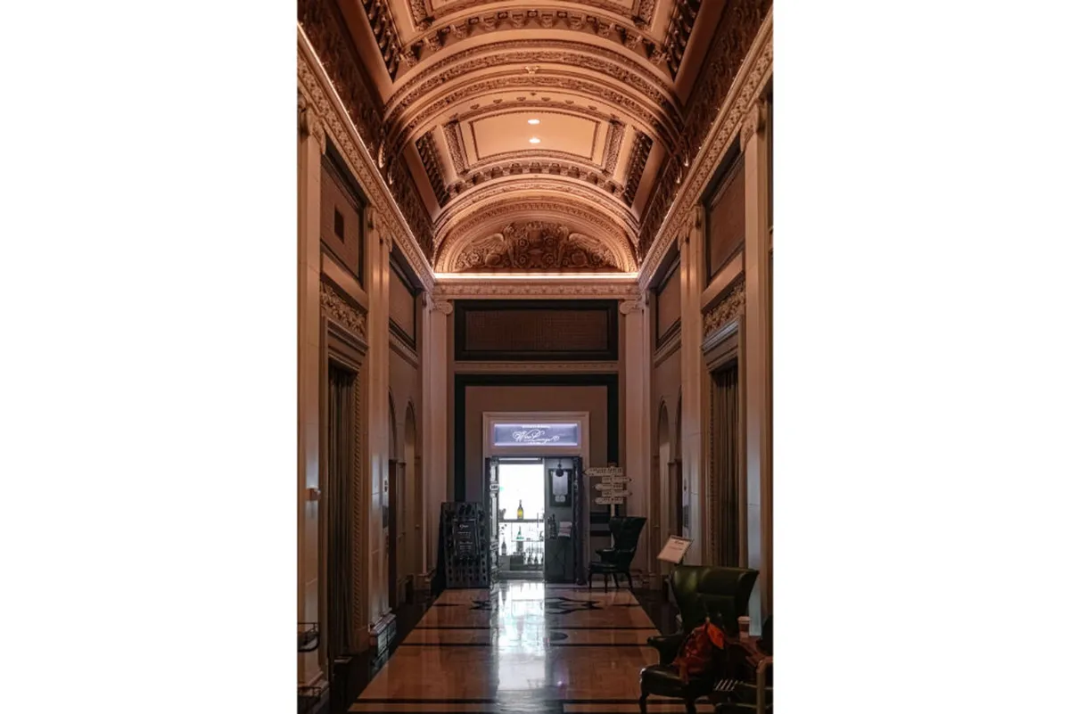 This image features a sophisticated interior space with an ornate ceiling, intricate moldings, a polished floor, and a prominent doorway that leads directly outside. The grand entrance creates a transitional feeling between the interior and the vibrant city beyond, evoking the sense of stepping out to explore.