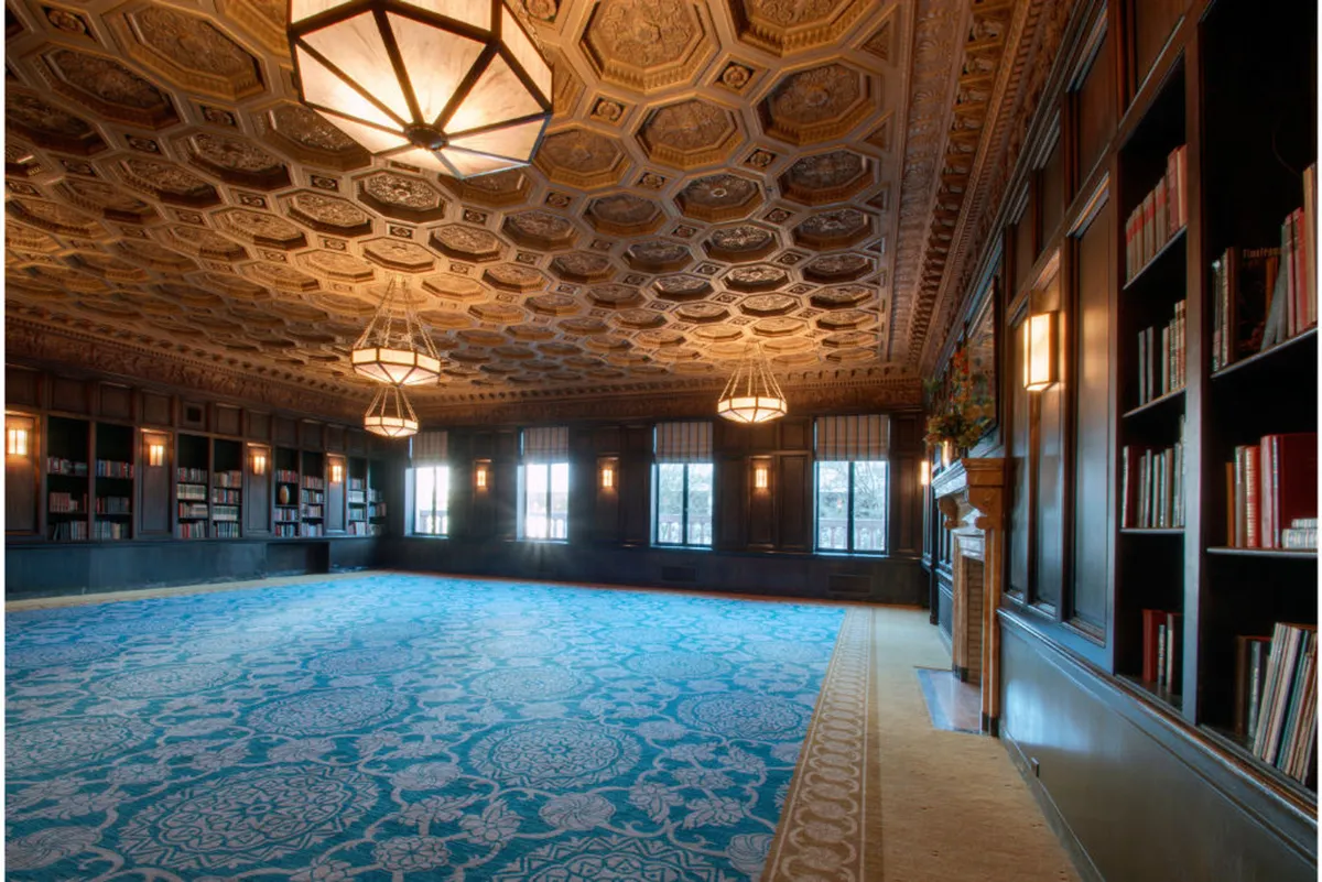 A tranquil, sophisticated room with an ornate ceiling, blue carpet, tall bookcases, and generous natural light from large windows. The space feels both grand and approachable, ideal for quiet time or socializing in a pet-welcoming environment.