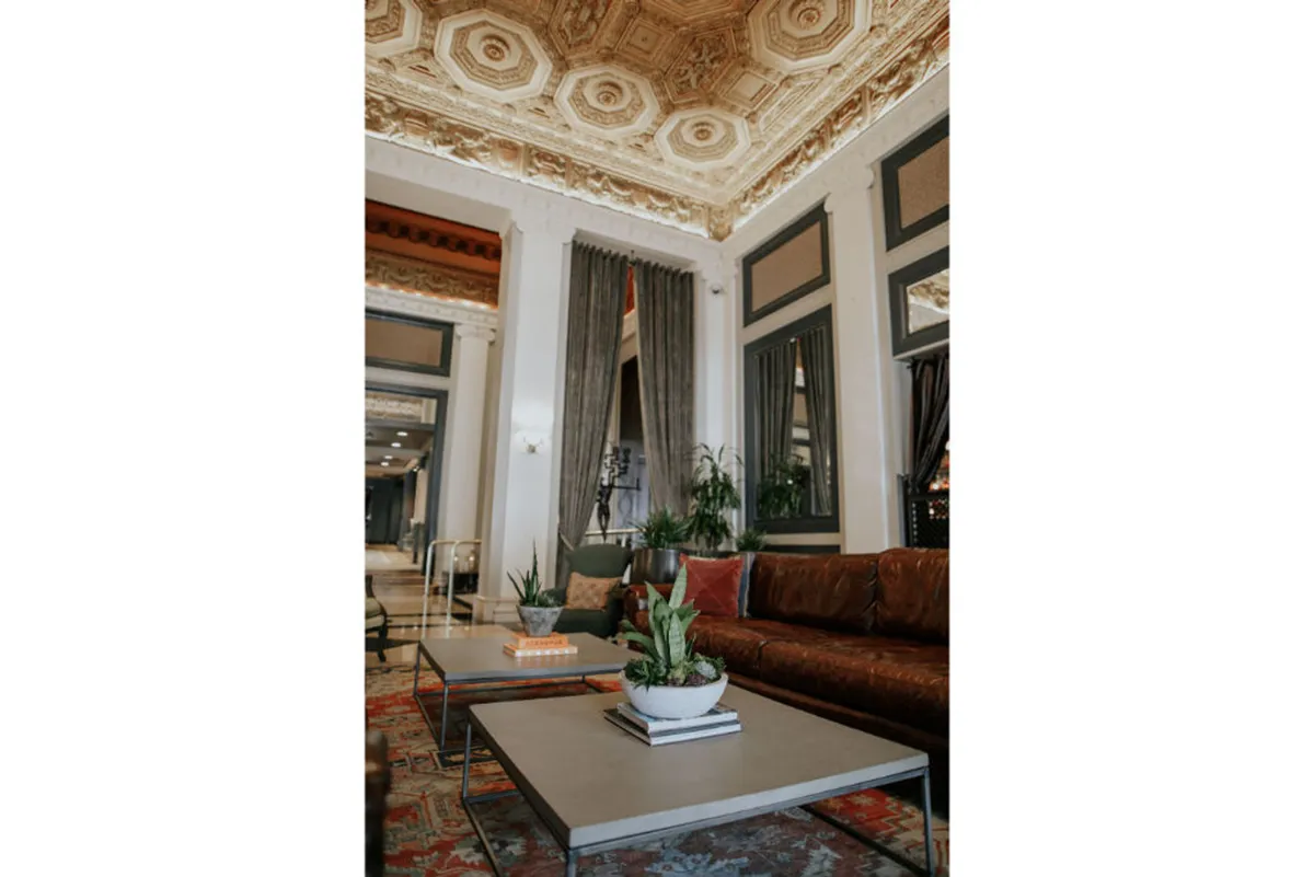 A welcoming lounge space featuring a comfortable sofa, coffee tables, and decorative plants. The room is warmed by natural light and ornate ceiling details, providing a sophisticated yet cozy environment ideal for relaxation or socializing.
