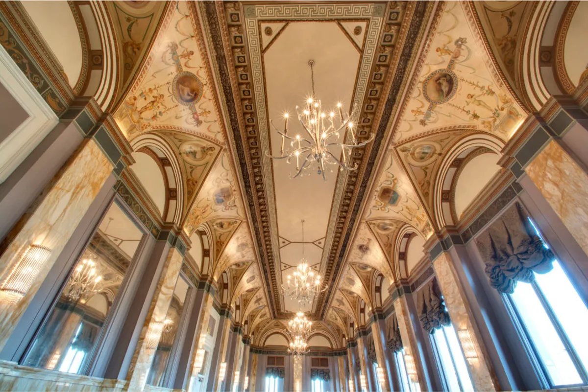 An opulent hall with ornate chandeliers, intricate ceiling designs, marble columns, and large windows with drapery. The grand entrance and windows provide captivating views, while the luxurious ambiance hints at gatherings, art exhibitions, or celebrations.