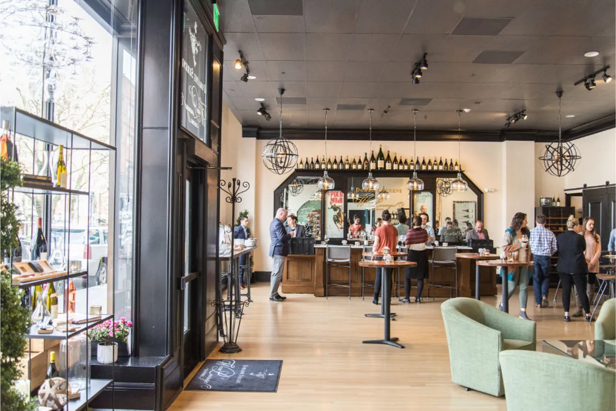 This lively interior image centers on a bar counter surrounded by hanging light fixtures, a glass display filled with wine bottles, and a comfortable seating area. The scene captures the energy of social gatherings and wine tastings in an inviting communal atmosphere.