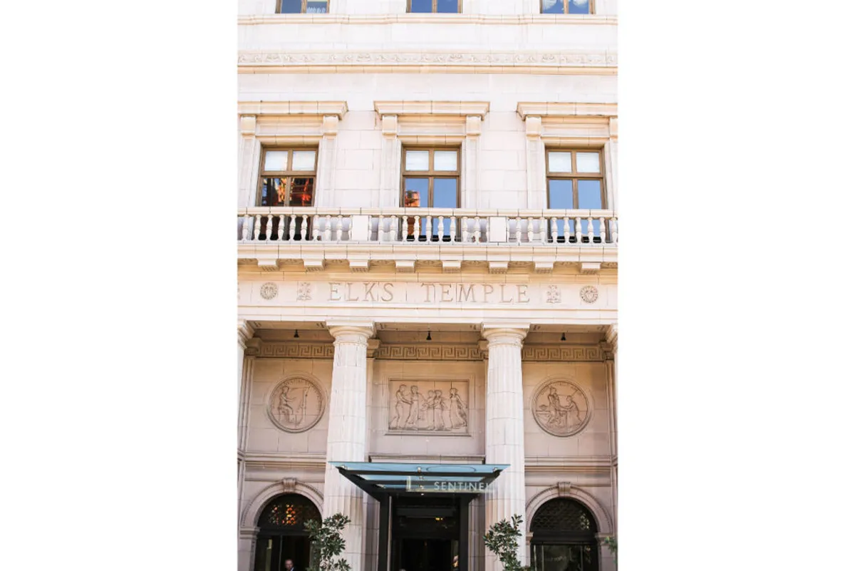 A detailed look at the Elks Temple sign, accompanied by ornate columns, decorative reliefs, and large windows, suggesting a blend of historic elegance and contemporary exploration. The mood is elegant and welcoming, inviting engagement with the area’s cultural history.