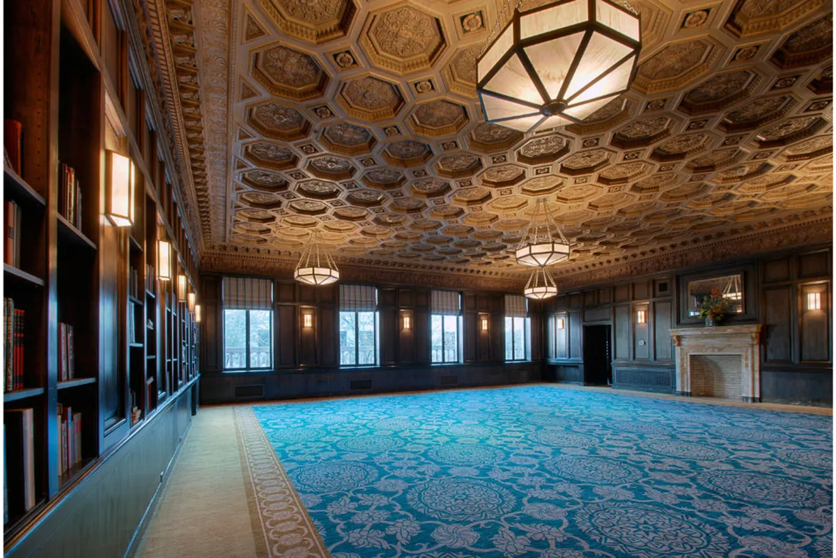 A grand room with an ornate ceiling, tall bookshelves, a cozy fireplace, and large windows that offer captivating views. The decorative carpet and historical decor create a warm, welcoming environment ideal for unwinding or socializing after a day out.